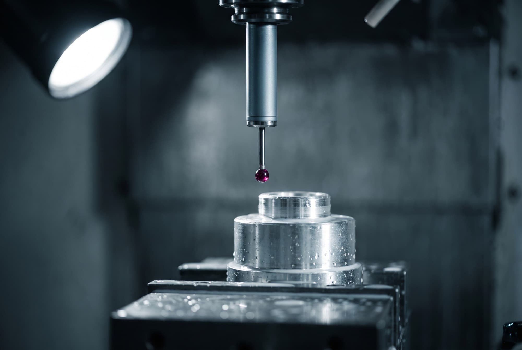 A spindle-mounted touch probe with a ruby stylus poised over the top face of a stepped cylindrical aluminum part, the part seated on a rectangular fixture block, coolant still beaded on the machined surfaces.